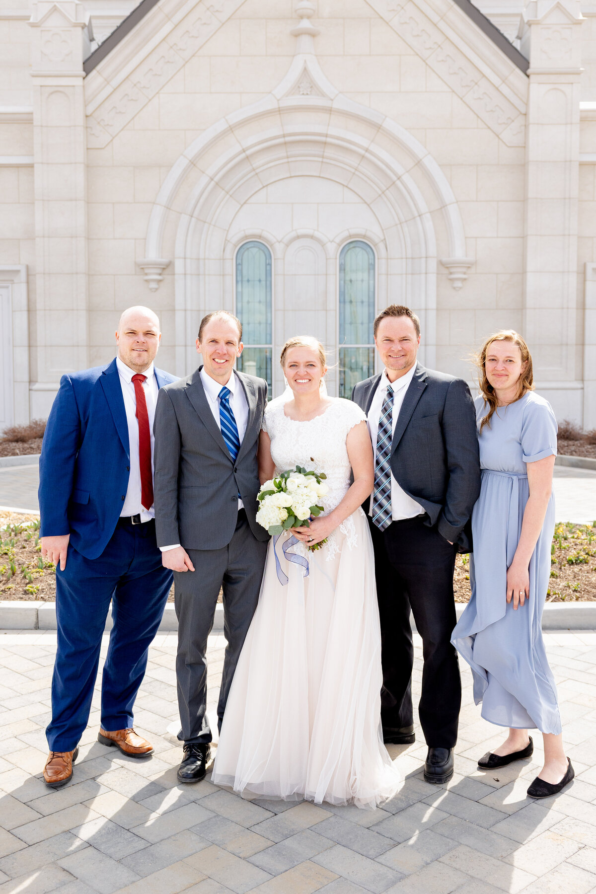 Bride smiles with five family members in formal attire, including suits and dresses, posing in front of the Taylorsville Utah LDS Temple after the sealing ceremony.