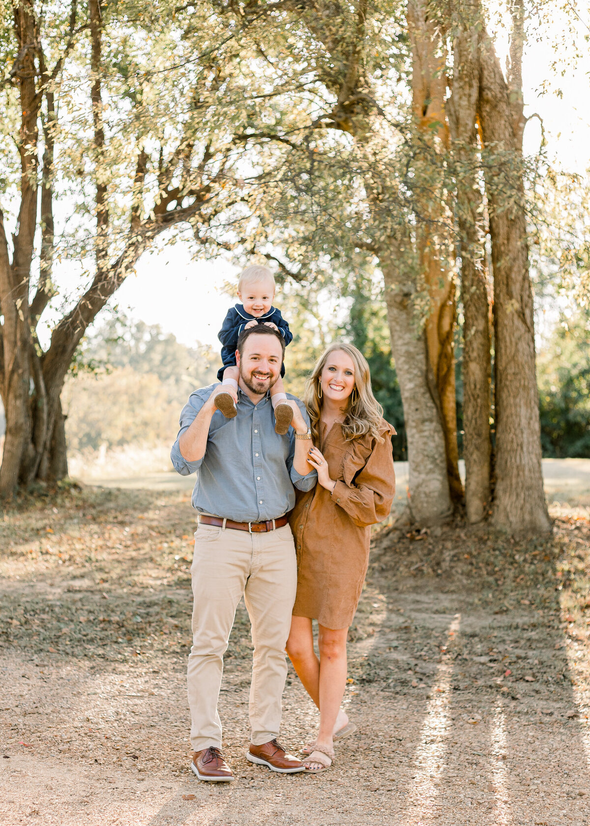 Outdoor family session with mom, dad, and toddler boy. Toddler boy is in navy outfit on dad's shoulders. Beautiful light shines through the trees behind them.  Image captured by Brandon, MS Family Photographer.