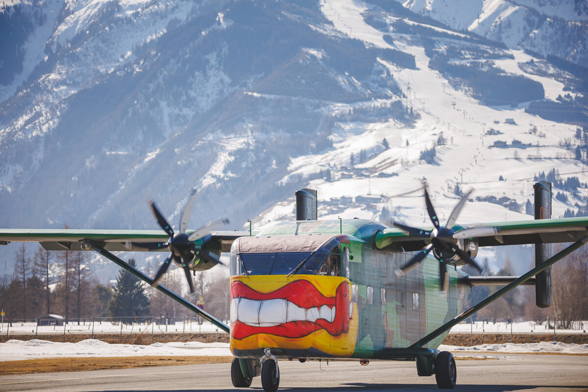 Die Pink Skyvan  steht am Boden. Dahinter sind verschneite Berge zu sehen.. Zu sehen ist das Flugzeug . Die bunte und auffällige Lackierung sticht heraus