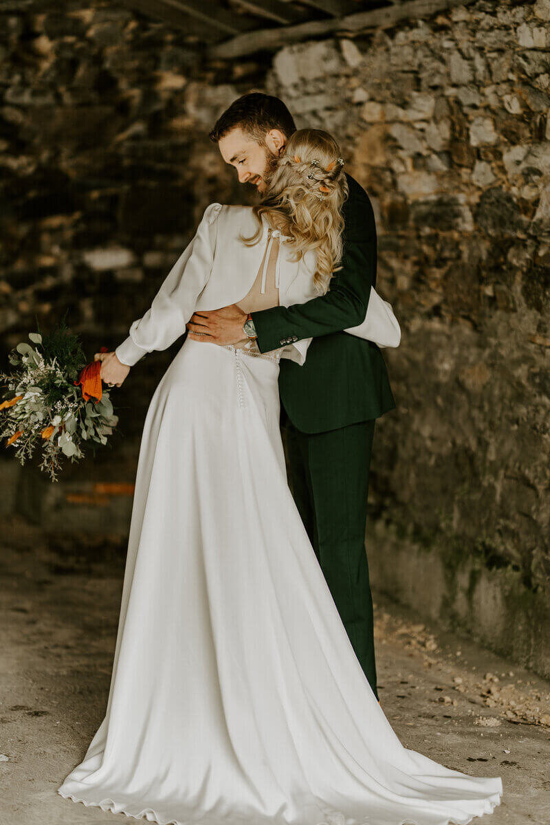 Mariée de dos, bouquet à la main enlaçant son mari dans un décor de vieilles pierres sous l'objectif de Laura, photographe professionnelle en Vendée.