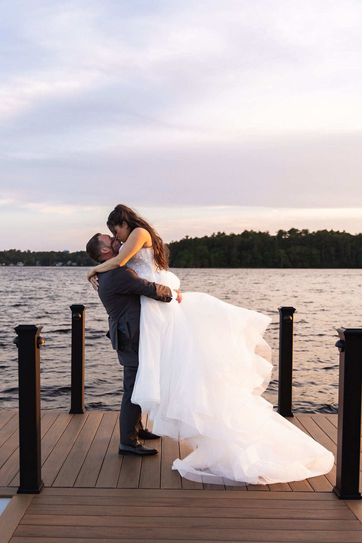 The Lakehouse Inn MA  | Kelsey Sheehan Photography Timeless Rhode Island Weddings | A bride in a flowing white gown is joyfully lifted by a groom in a suit on a lakeside dock, with a serene water and forest backdrop under a soft, pastel sky.