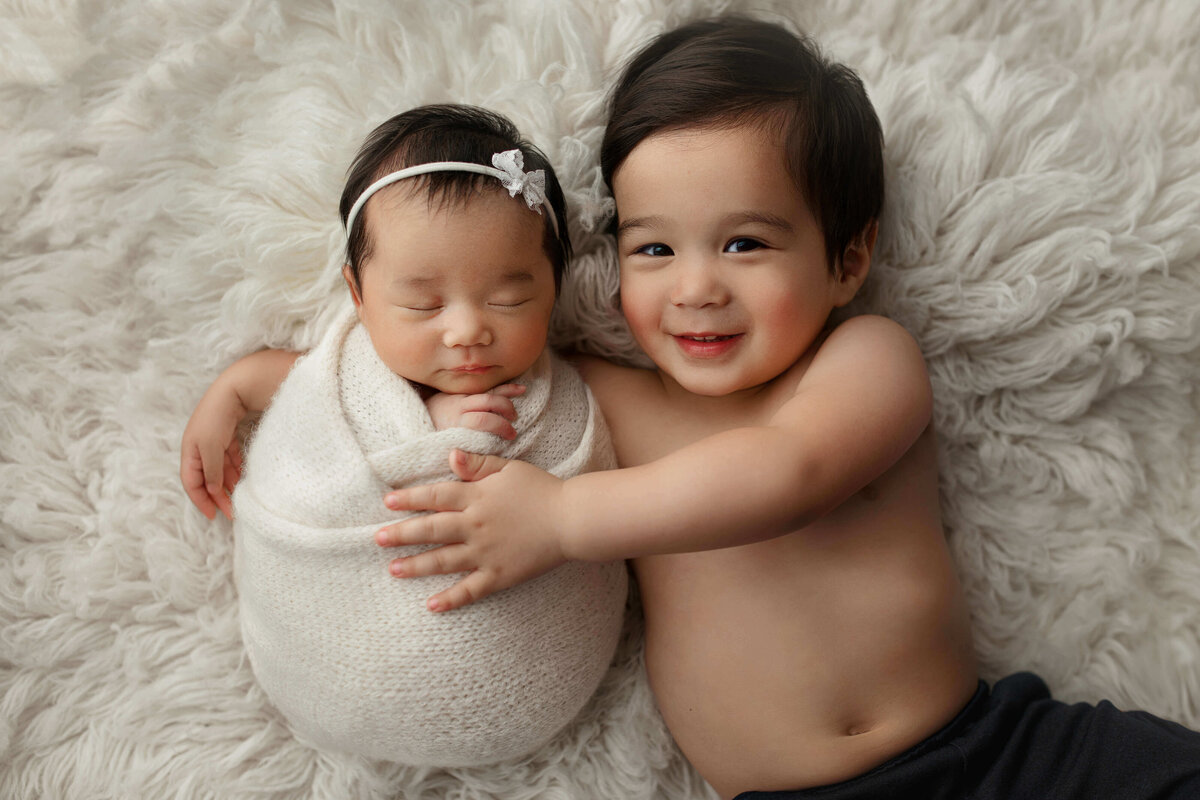 Newborn girl in a white wrap with bow laying with her big brother on a  white fur rug at a baby photo shoot in Northern VA