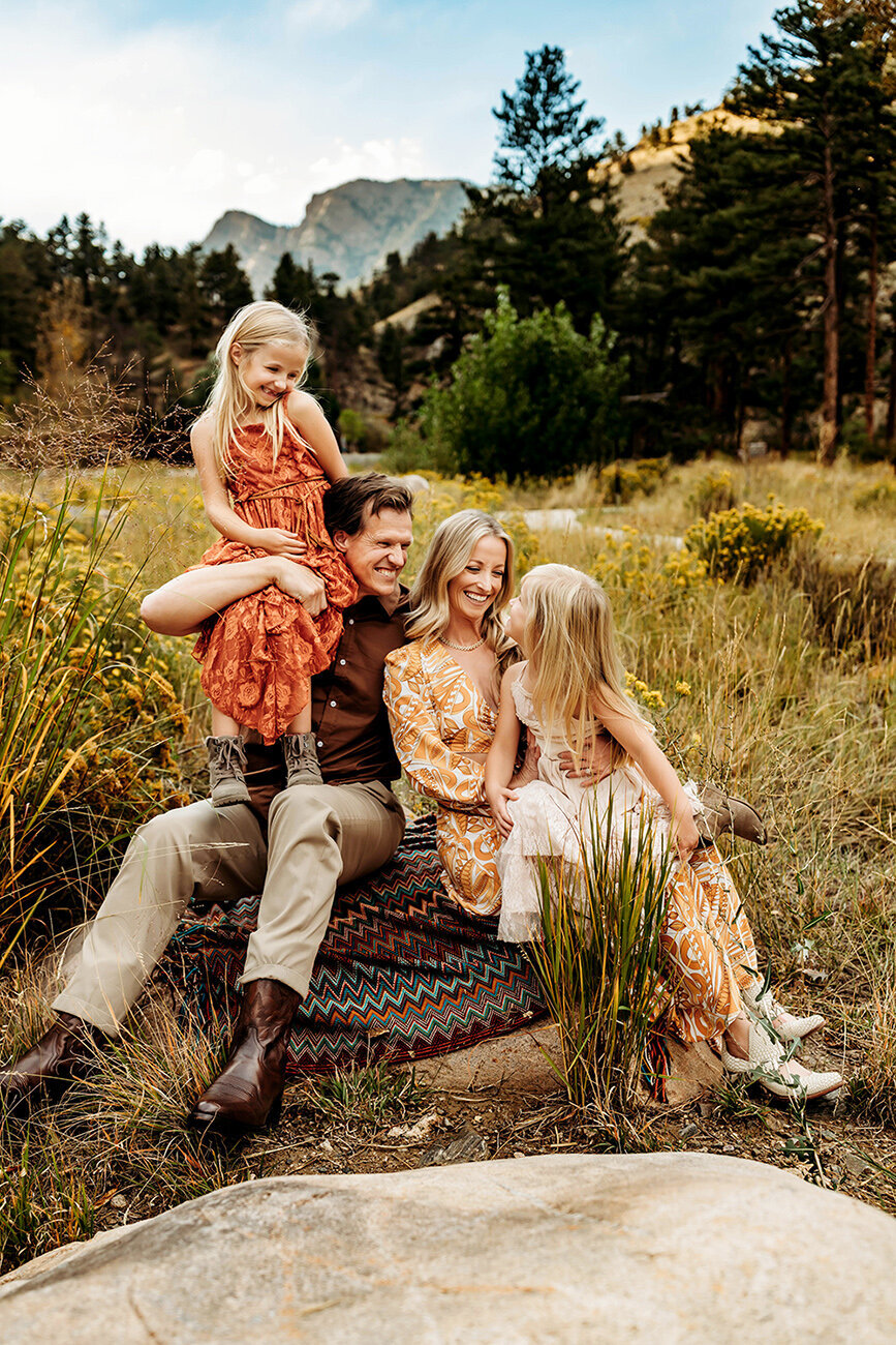Dad, mom and 2 school aged blond daughters laughing together in the Estes Park mountains.