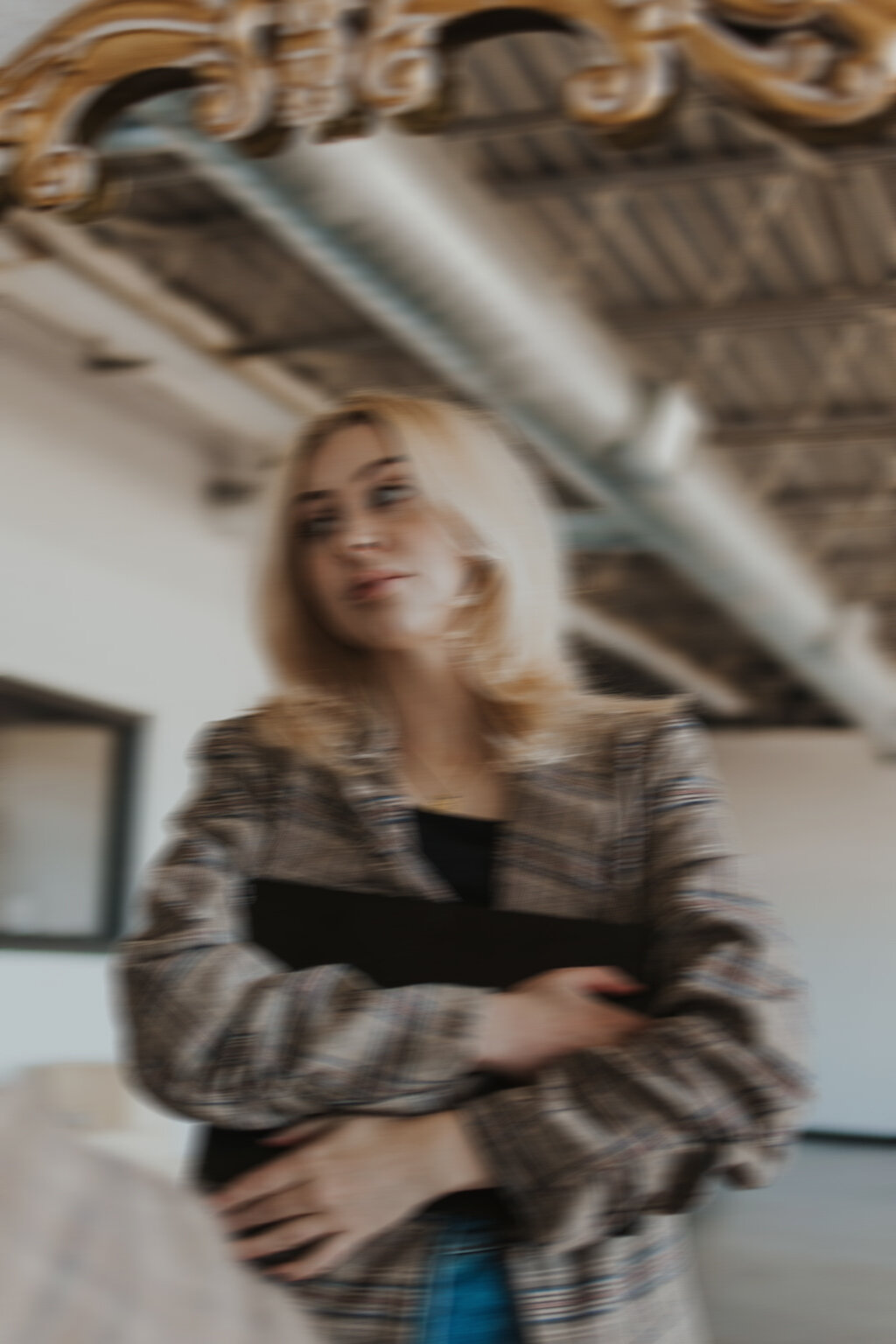 A blurry image of a woman, wearing a brown, plaid suit jacket, holding a black laptop.