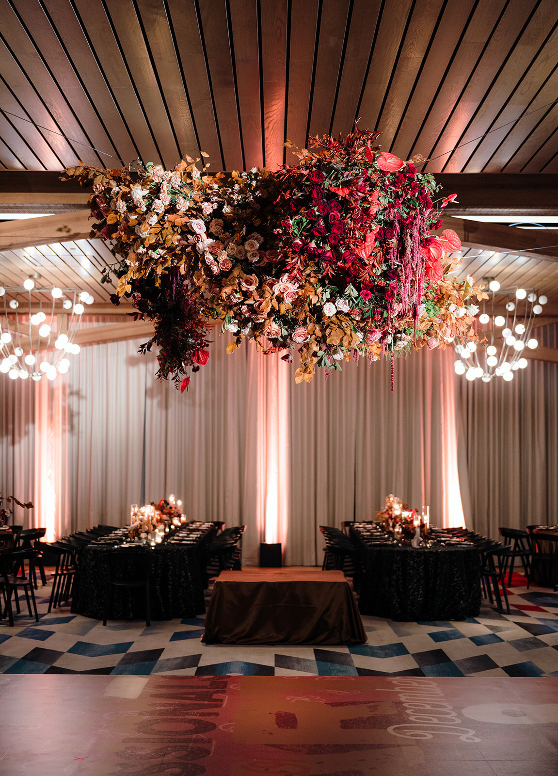 Large floral hanging installation decorates dance floor of reception space. Dark and moody event design with monochromatic red, burgundy, copper, cream, and blush. Sculptural and product clustered floral design. Interacting floral clouds for winter wedding industry event. WIPA Nashville Gala 2024. Floral design featuring orchids, anthurium, and roses. Design by Rosemary & Finch Floral Design in Nashville, TN.