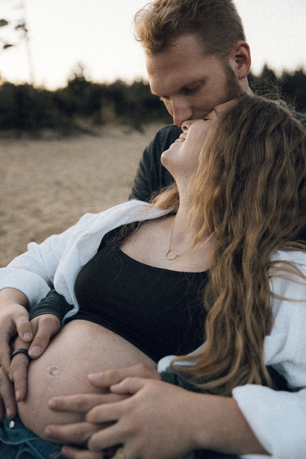 Woman leans back into man while man holds her pregnant belly during a maternity photo session on the beach in Oregon. 