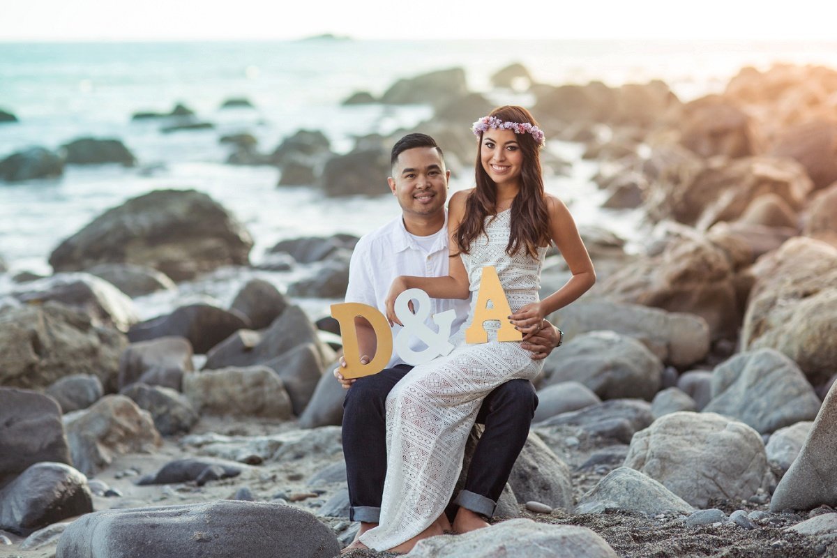 Bride to be sits on her fiance's lap holding their initials while sitting on rocks at the beach for photo shoot