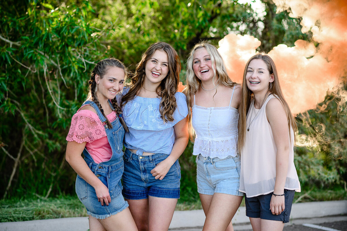 4 girls in summer outfits standing laughing with their arms around each other with an orange smoke bomb in the background while posing for a senior picture photographers near me