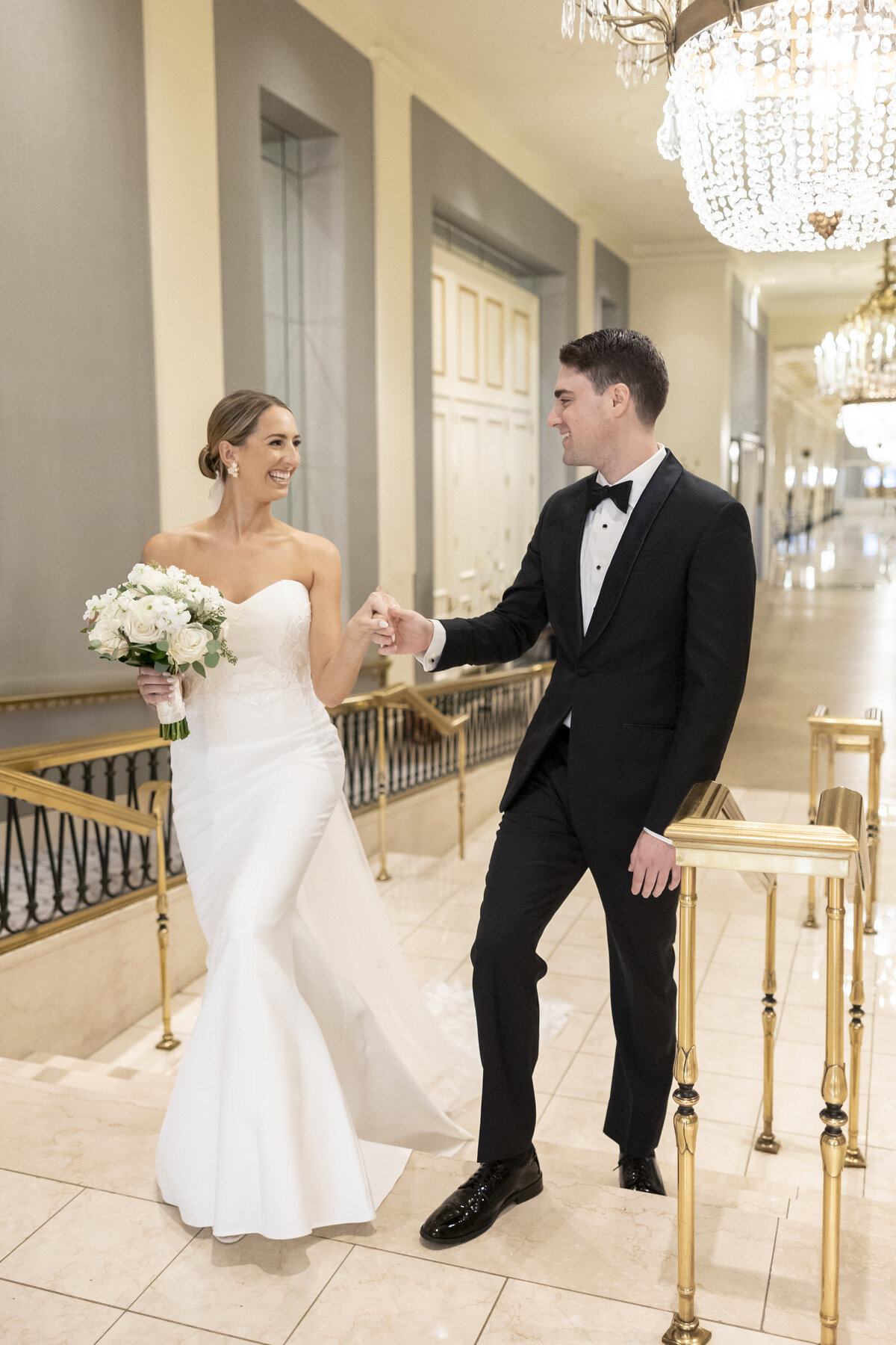 A bride and groom walk down a staircase in an elegant setting. The bride is in a white gown holding a bouquet, while the groom is in a black tuxedo. They are smiling at each other, with chandeliers and a long corridor in the background.