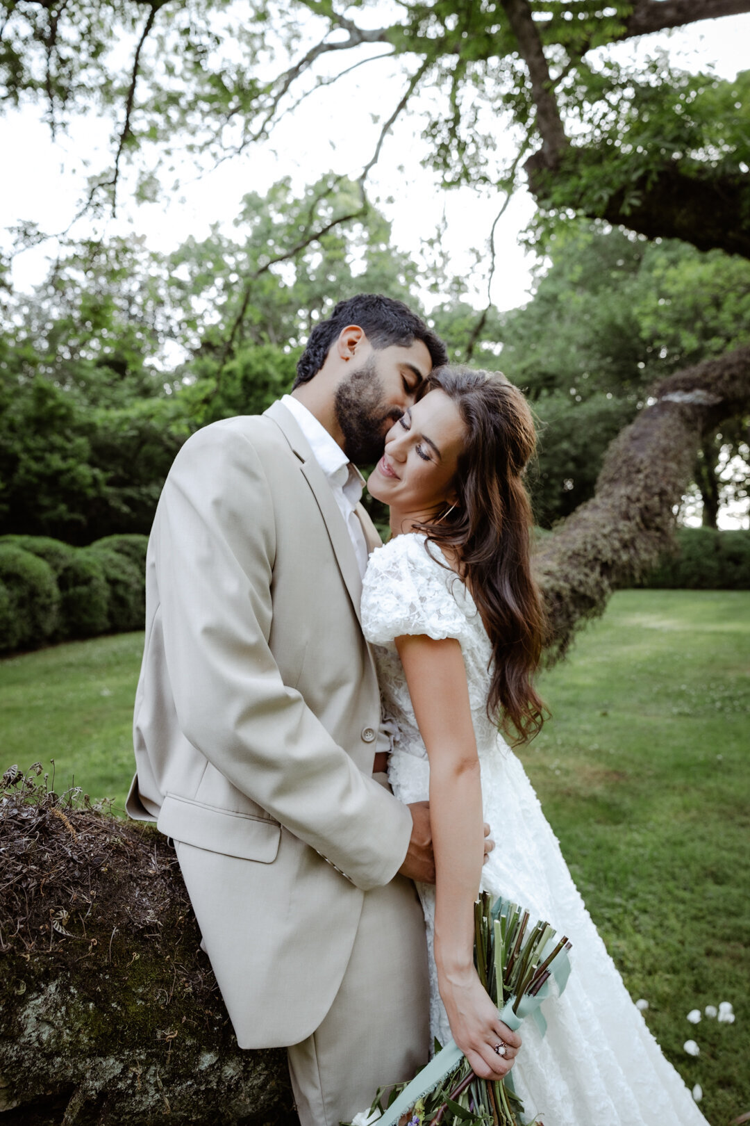 Groom sitting on a tree branch while bride rests her head on his shoulder, holding a vibrant bouquet.