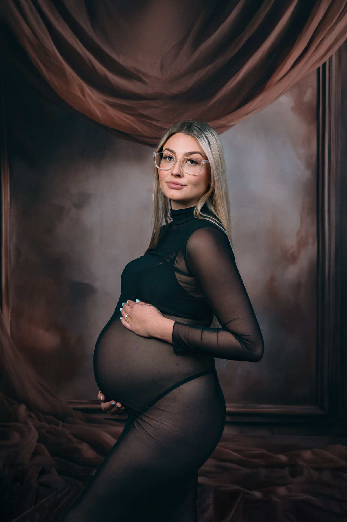 brown toned draped fabric behind a stunning blonde pregnant lady in sheer black high neck dress