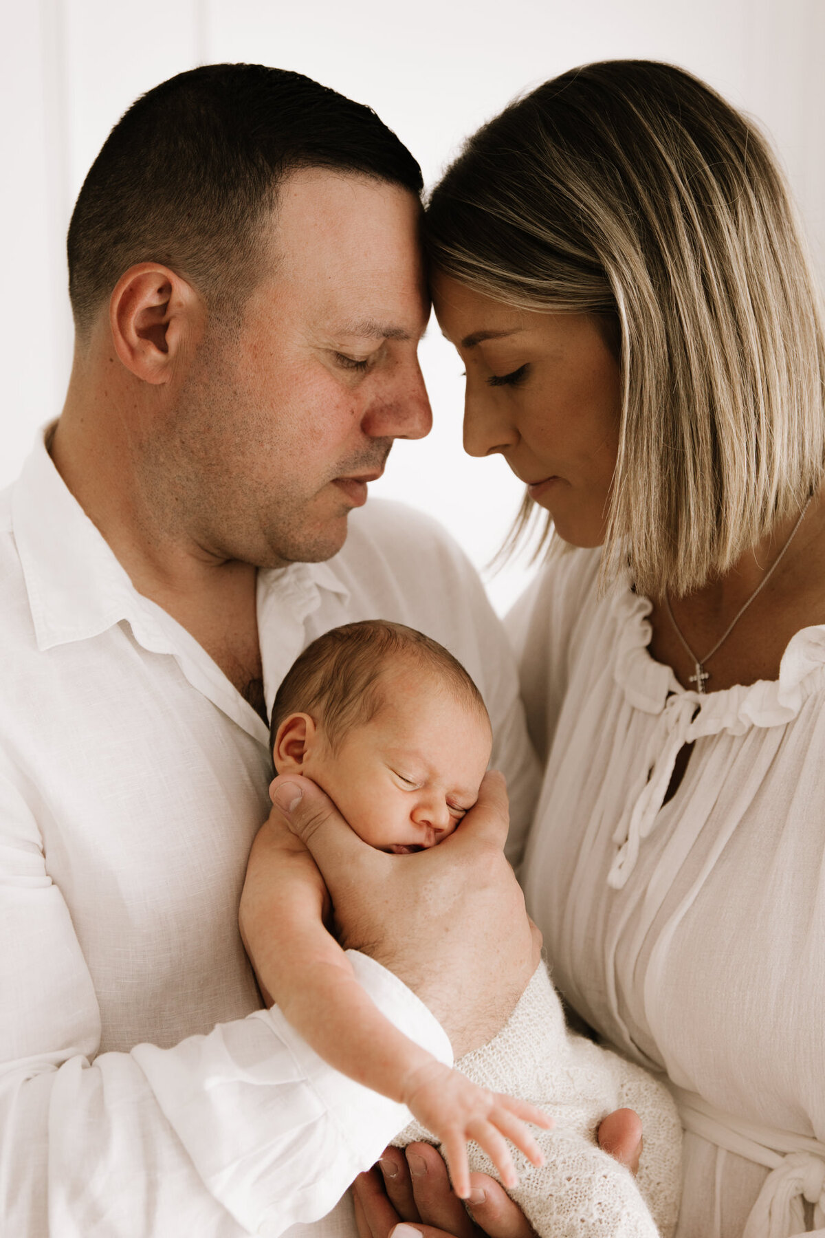 Mother and father gently cradling their sleeping newborn, foreheads touching in a quiet, connected moment during a newborn session in a softly lit Sydney studio.