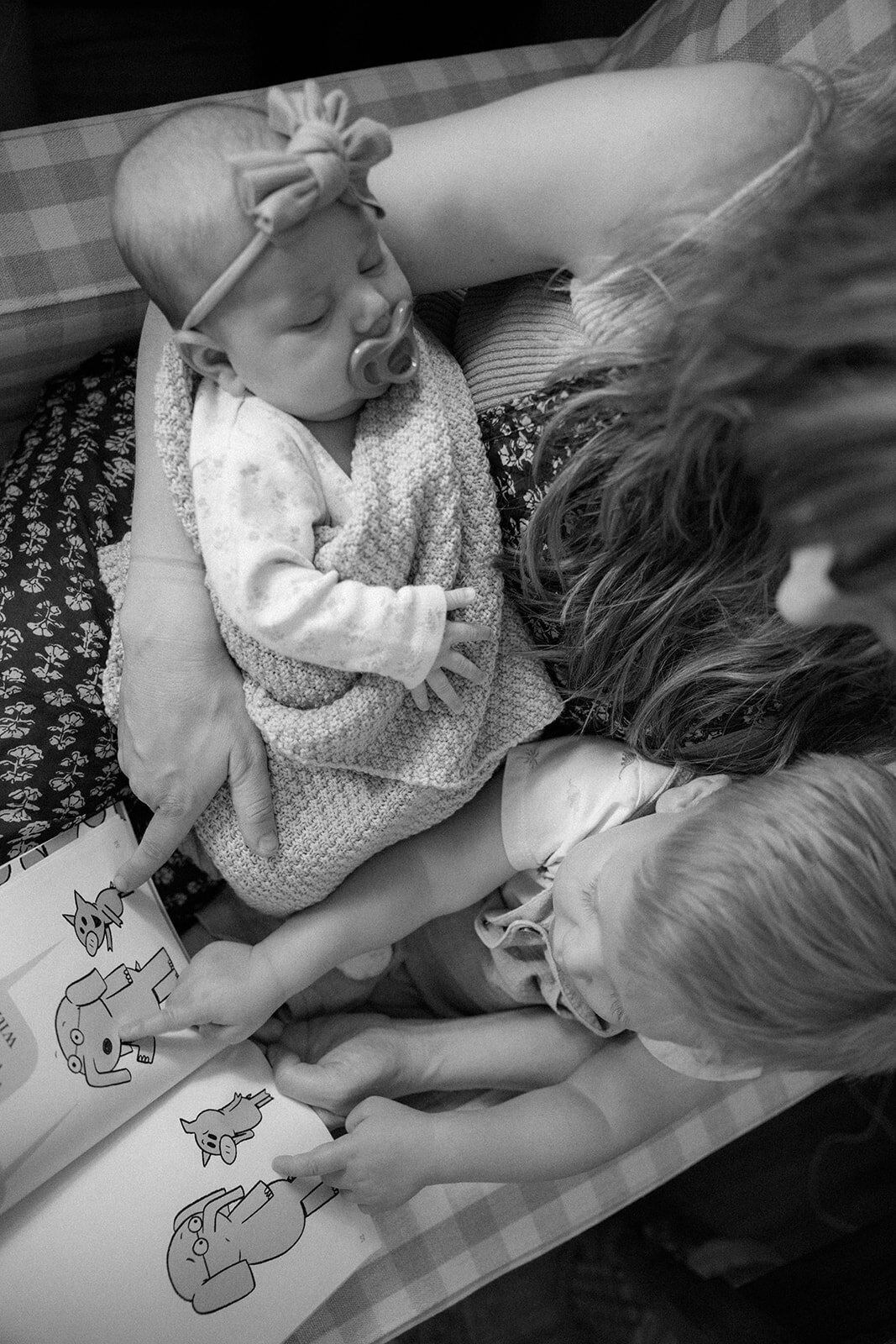 Newborn and toddler snuggle in their mom's lap while she reads a children's book to them. 