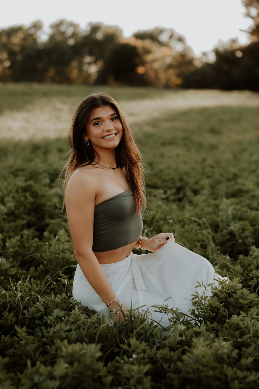A young woman with long hair, wearing a green crop top and white skirt, sits on a field of green foliage. As she smiles at the camera, captured by a New England Family Photographer, trees and a soft, golden sky create a perfect backdrop.
