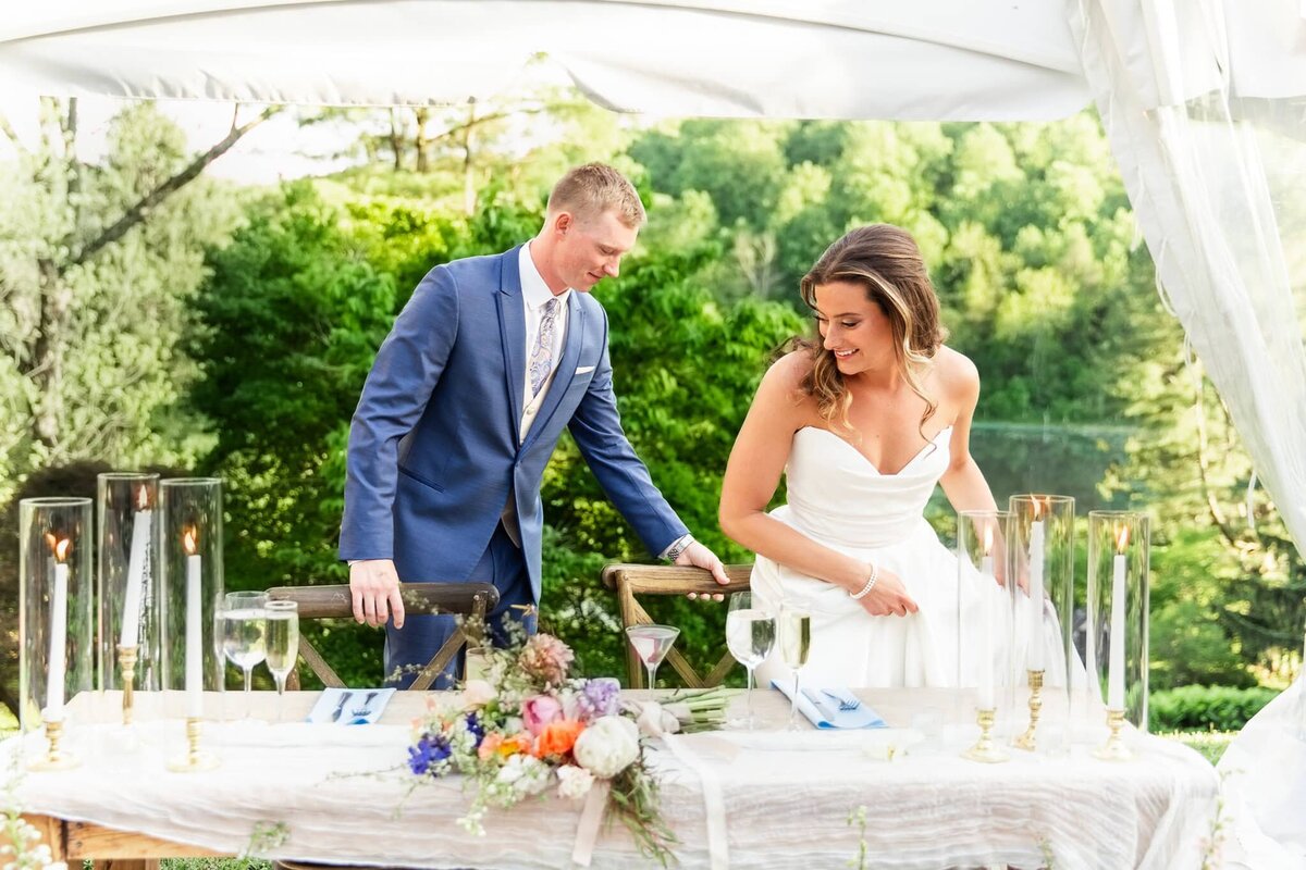 A bride and groom take their seats at a sweetheart table during an outdoor wedding reception just an hour from Lancaster, PA. The bride, in a strapless gown, smiles as the groom helps pull out her chair. The table is adorned with tall glass candle holders, champagne flutes, and a colorful floral arrangement of orange, purple, and white blooms, set against a lush green backdrop.