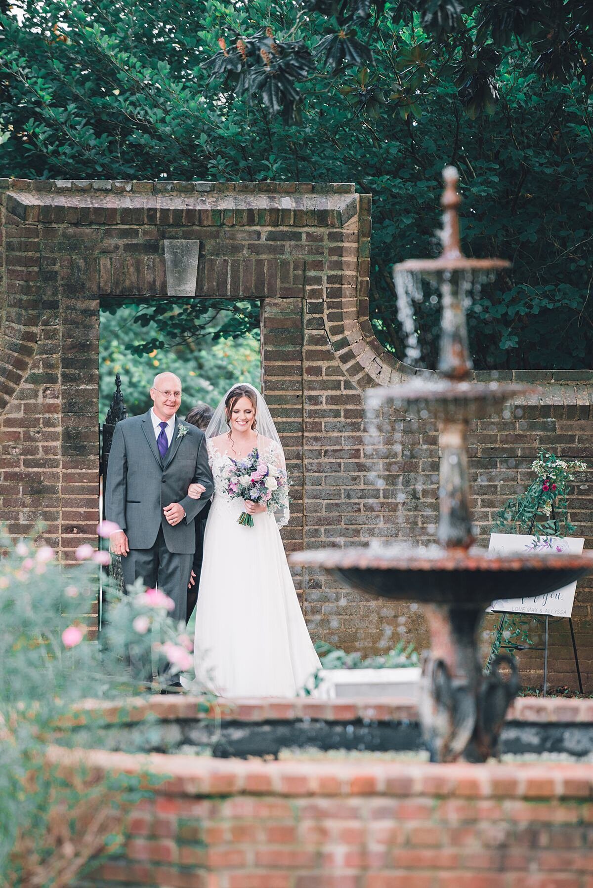 A bride wearing a strapless wedding dress and long veil walks arm in arm  with her father holding a large pink and purple bouquet as they walk through the brick courtyard garden at Rippavilla Plantation towards the three tiered fountain.