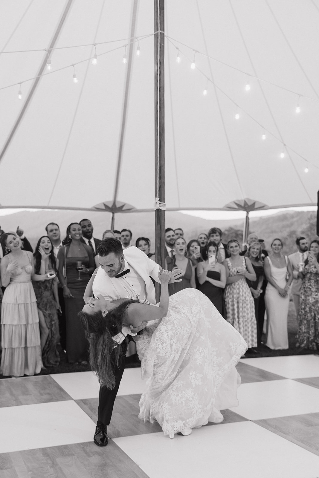 Black and white photo of the groom dipping the bride during their first dance as guests cheer under a tent in Highlands, North Carolina.