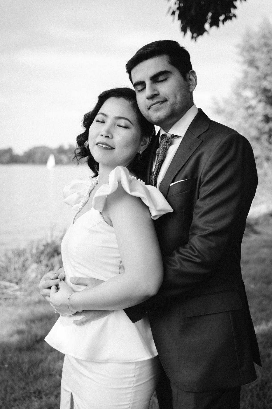  A black and white portrait of a couple standing close together outdoors. The man, in a suit, wraps his arms around the woman from behind. They both have their eyes closed with peaceful expressions. The woman, wearing a white dress with pearls, leans back into him. They are standing near a body of water with a blurred, light background.