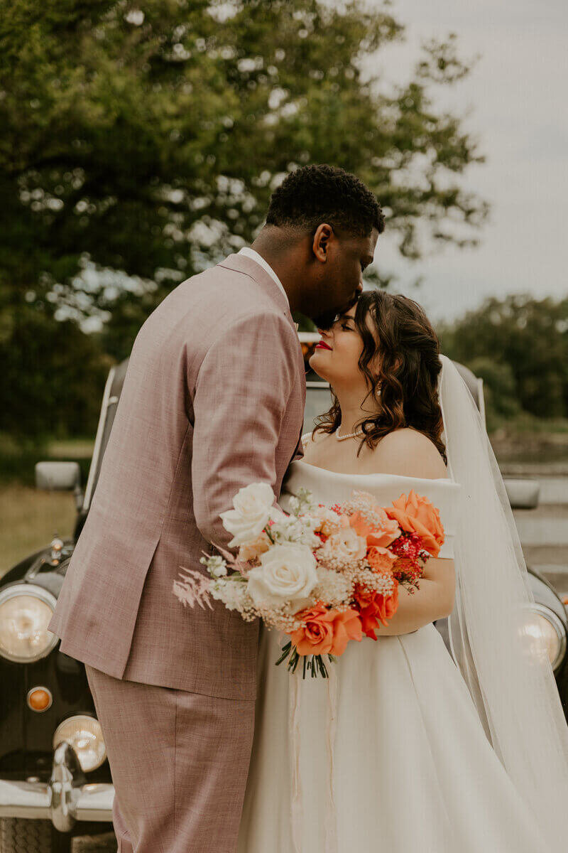 Marié en costume rose donnant un baiser sur le front de la mariée avec un bouquet de fleurs blanches et oranges. Voiture noire ancienne en arrière plan. Photo prise par Laura, photographe de mariage en vendée.