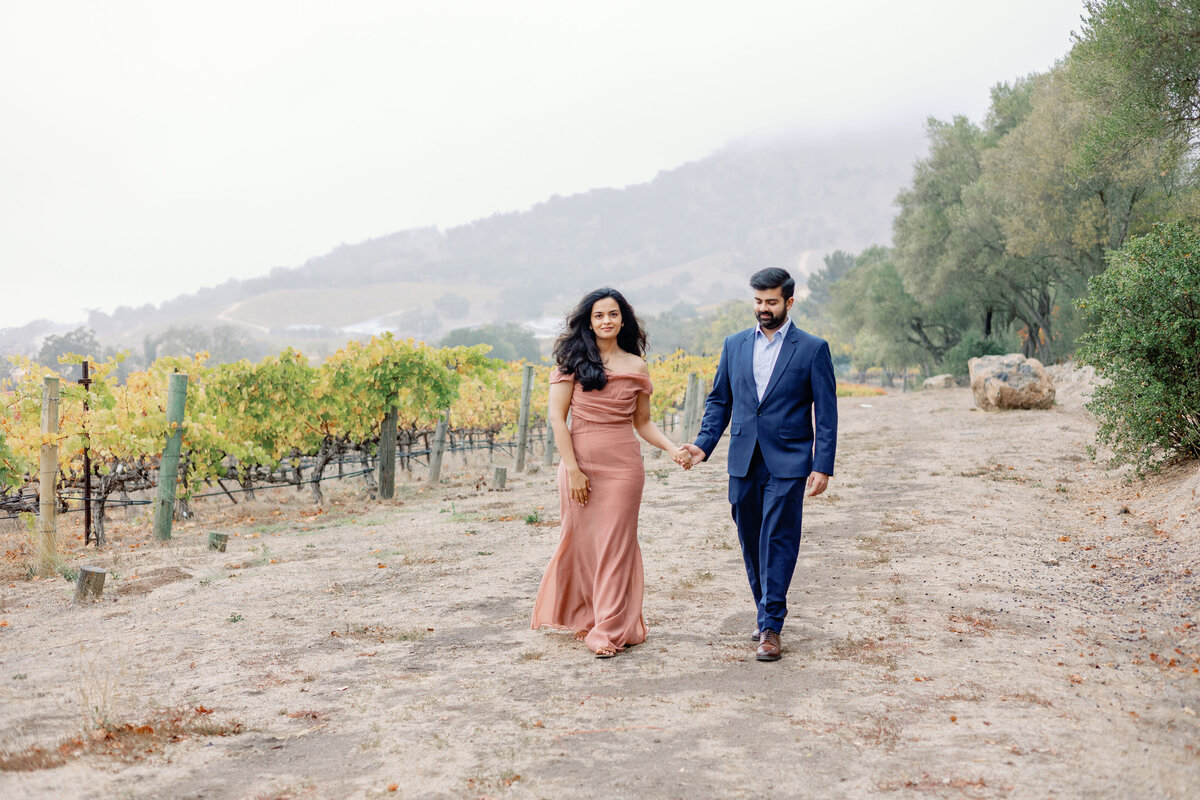 bay area photographers photographs vineyard engagement photos with man and woman holding hands and walking next to rows of grape vines in Napa