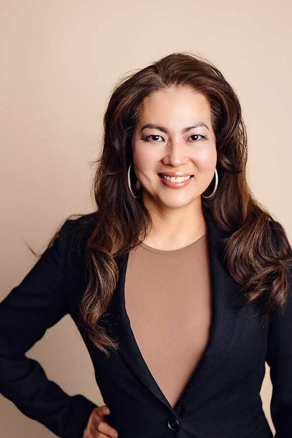 Woman in tan shirt and black jacket smiles while putting hand on hip.