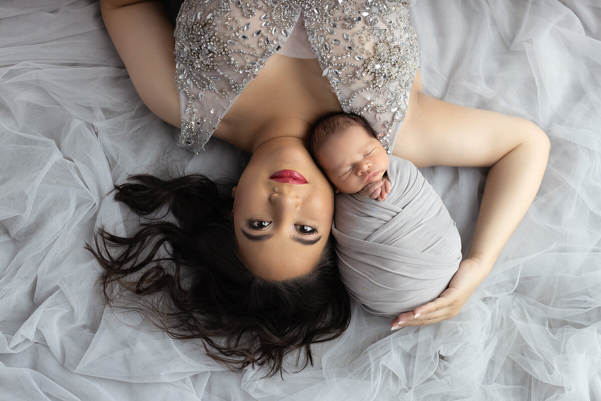 Elegant mother in a beaded gown lying on tulle with her swaddled baby beside her, featured in a Dallas newborn photography session.