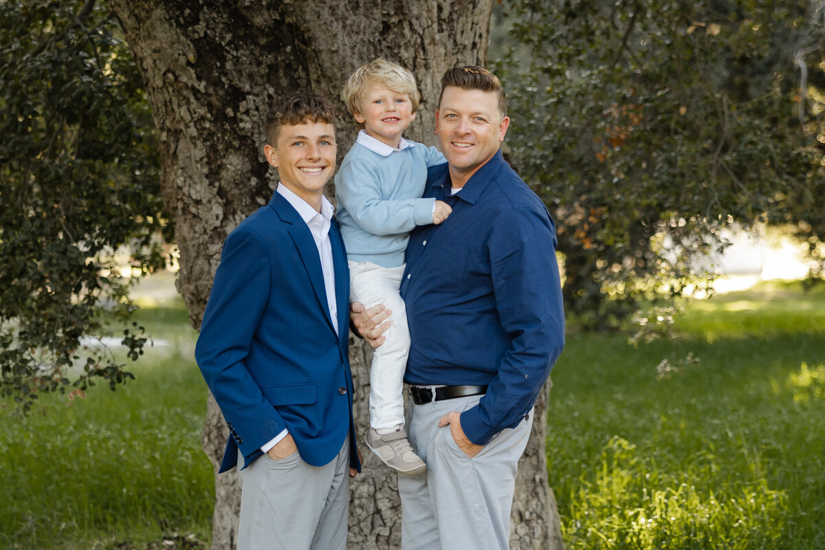 Father standing closely with his two sons, all dressed in soft blue and navy tones. Photographed by Yucaipa family photographer Kaitlyn Dawn Photography.