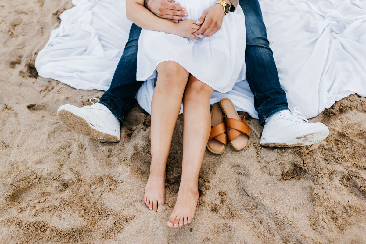 womans legs laid out in front of her as she sits in her fiances lap with the womans shoes to the left of her for beach engagement photos