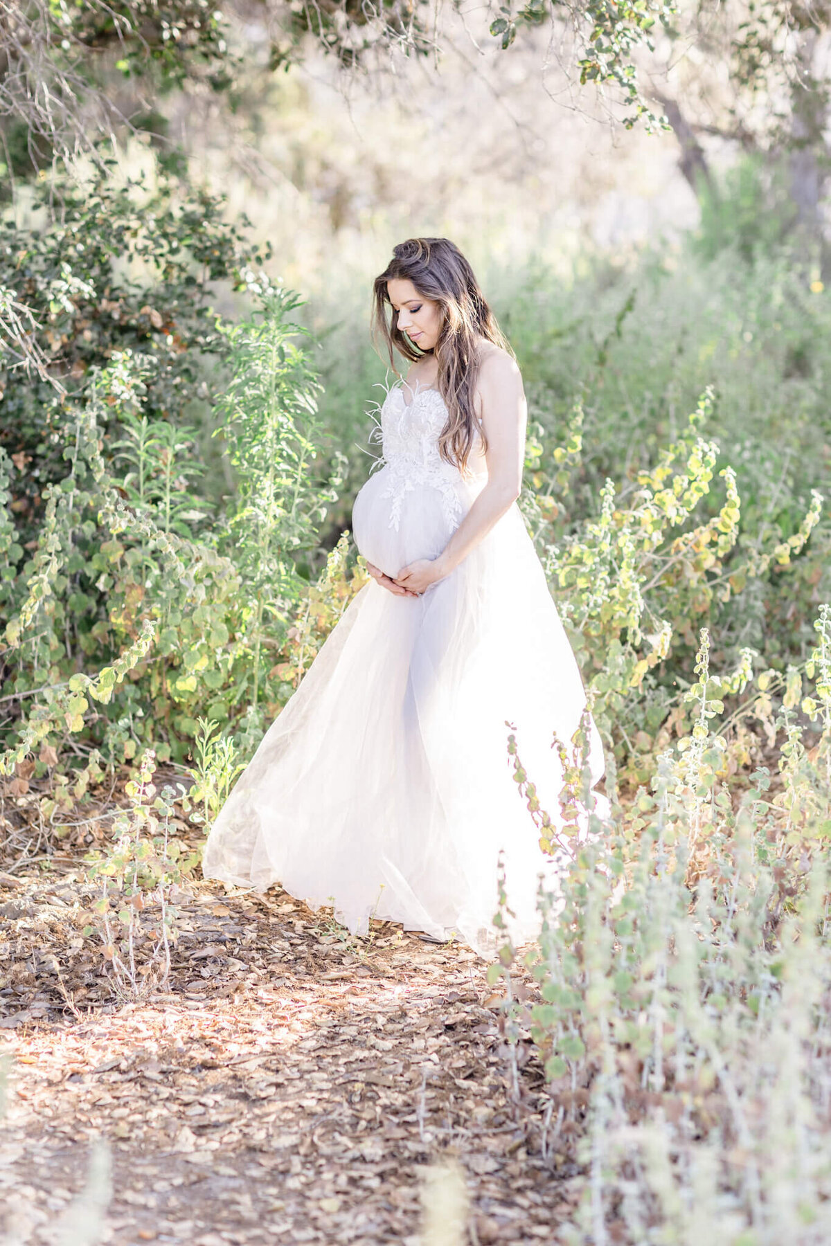 A pregnant woman in a long, flowing white dress stands outdoors in a sunlit, leafy setting. She gently holds her belly, surrounded by greenery and dappled sunlight, creating a serene and peaceful atmosphere.