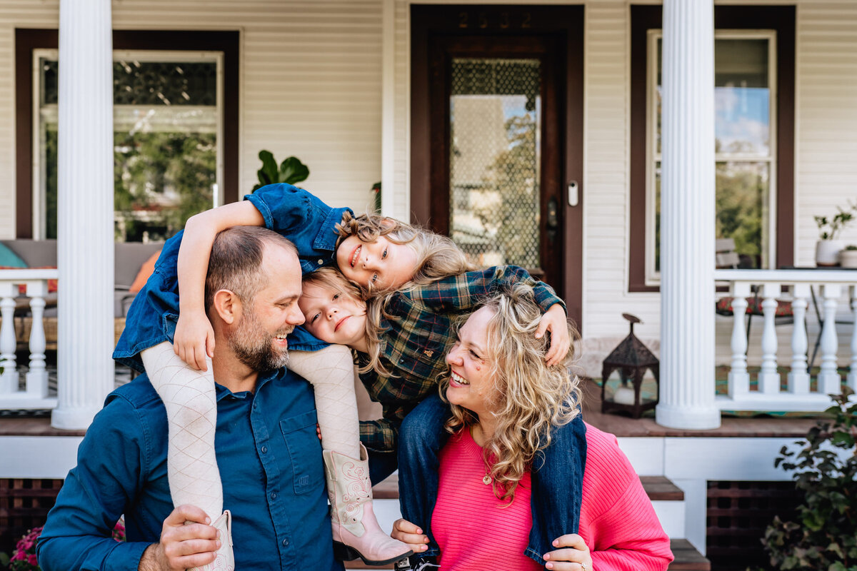 A joyful family moment on the front porch. The father holds one child on his shoulders while the child playfully leans on the mother’s head. The mother looks up and smiles warmly at her family.