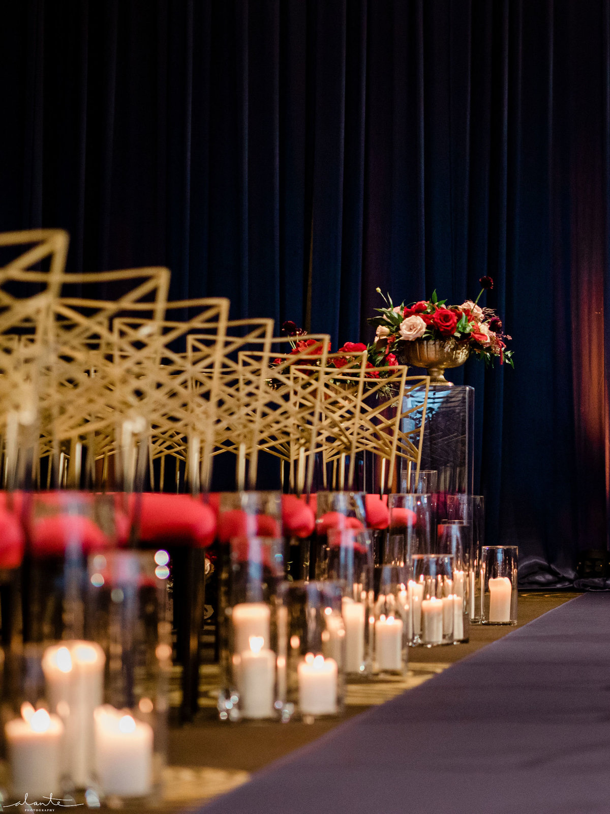wedding ceremony aisle decor with hurricane candles, blue aisle runner, and gold chairs with red seat cushions