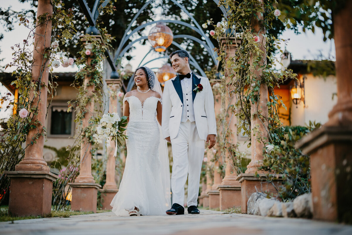 Bride and groom walking under the flowered pergola at Tenuta Corbinaia, elegant wedding in Tuscany.