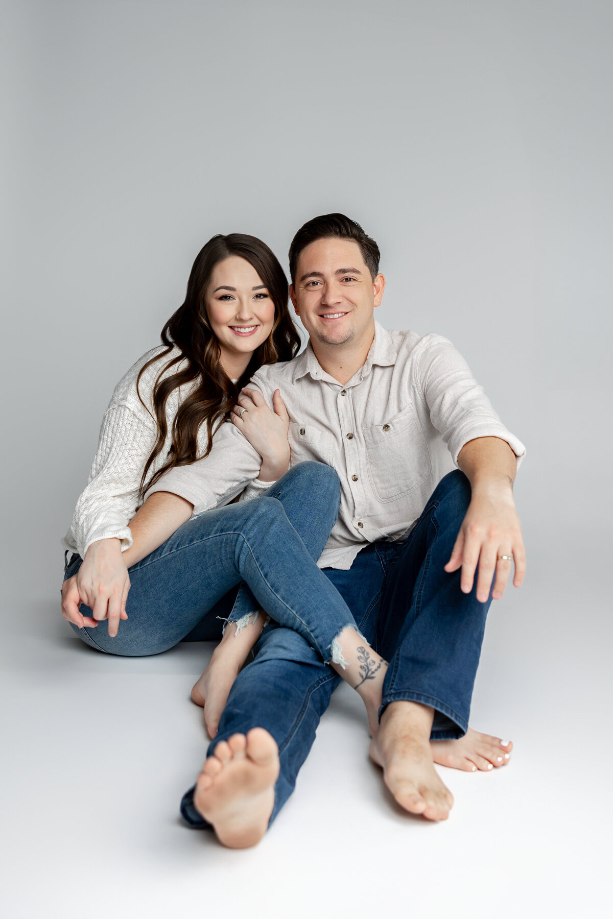 Husband and wife sitting together barefoot on studio floor, dressed in matching neutrals. Photographed by Yucaipa family photographer Kaitlyn Dawn Photography.