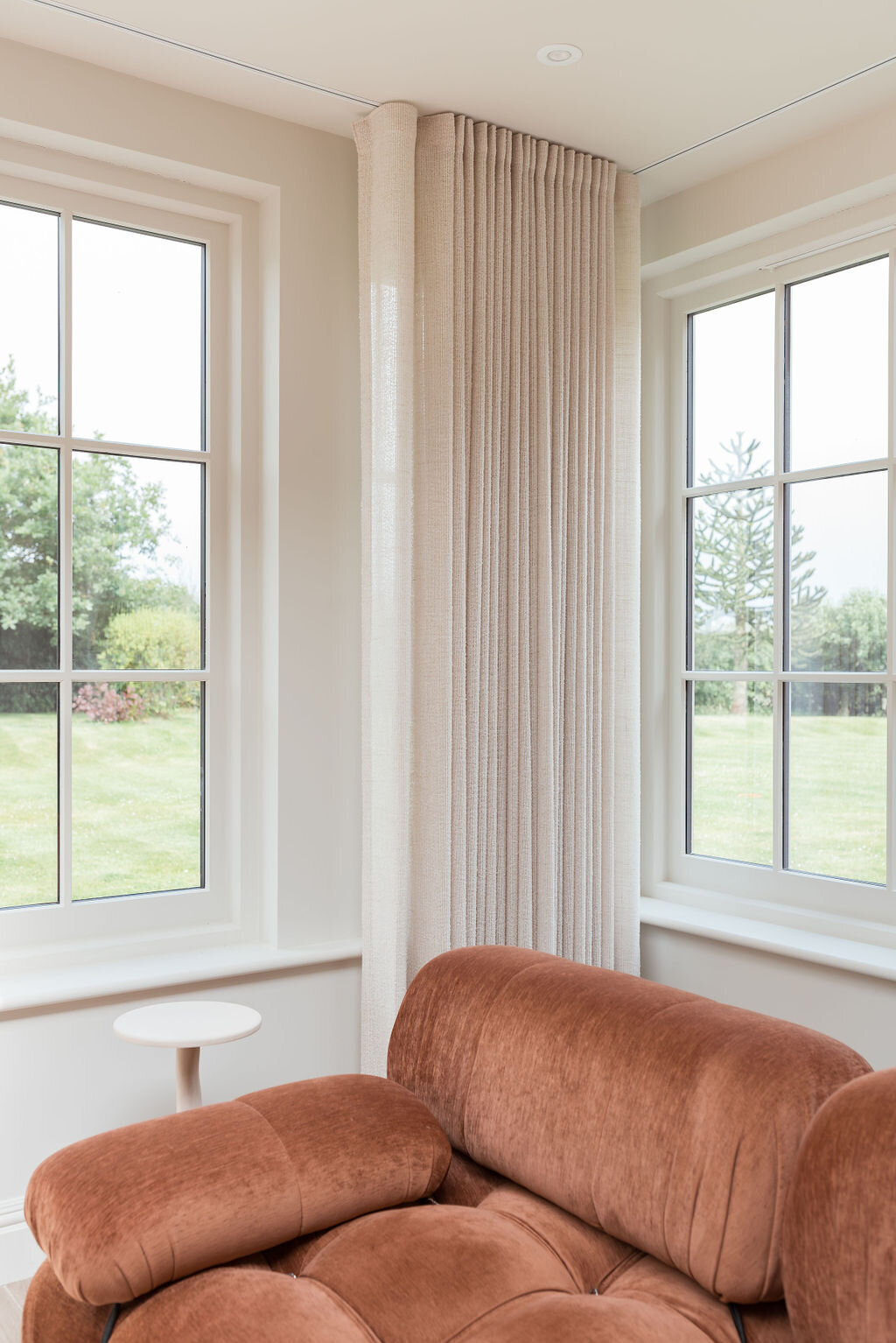 A rust orange, plush sofa with a small white side table, surrounded by natural light from large windows..