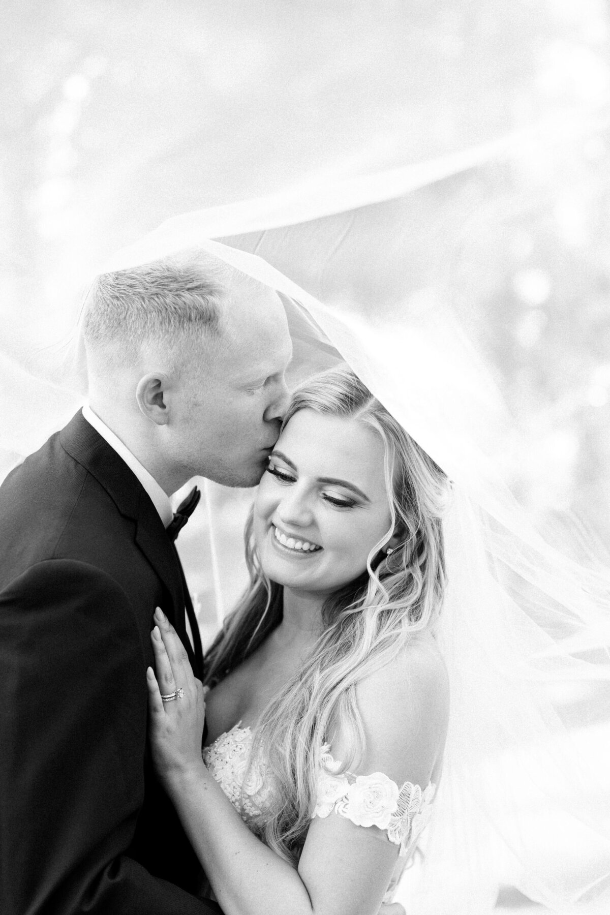 A groom kisses a smiling bride on the forehead under a veil in their wedding attire during their romantic wedding in Calgary at Norland Estate.