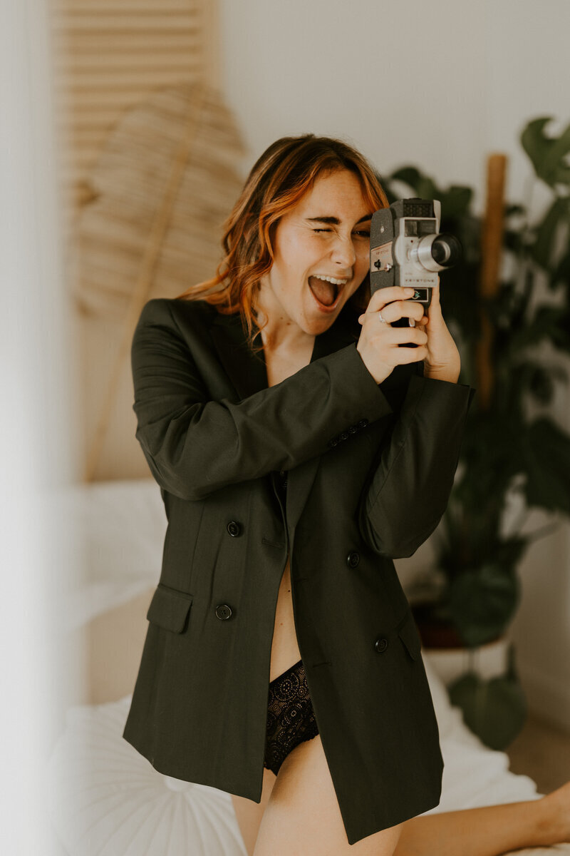 Femme rousse en lingerie, veste noire, s'amusant sur un lit, appareil photo vintage à la main, lors d'une séance photo portrait en Vendée.