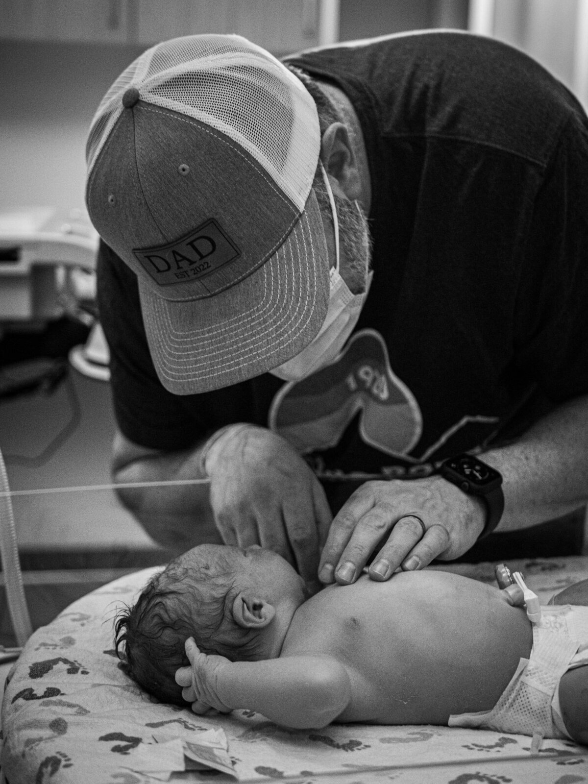 A new dad curiously inspects his newborn while it lays in a hospital warming bed.