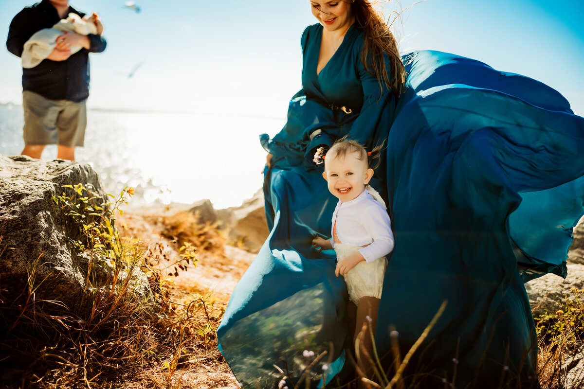 a women in a flowy blue gown on a super windy day playing with her toddler while her husband hold their newborn baby on the background