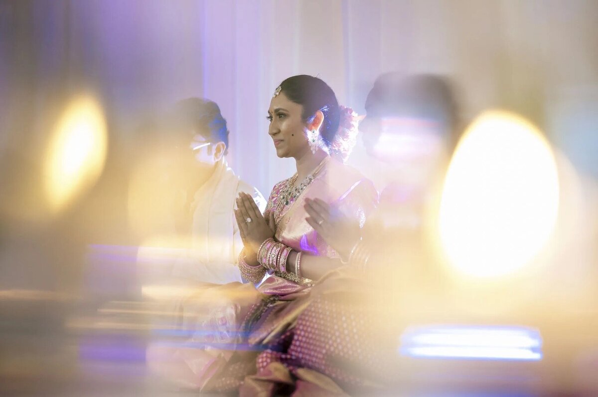A woman in traditional South Asian attire sitting with folded hands, ethereal with bokeh light surrounding her, reflecting cultural elegance.