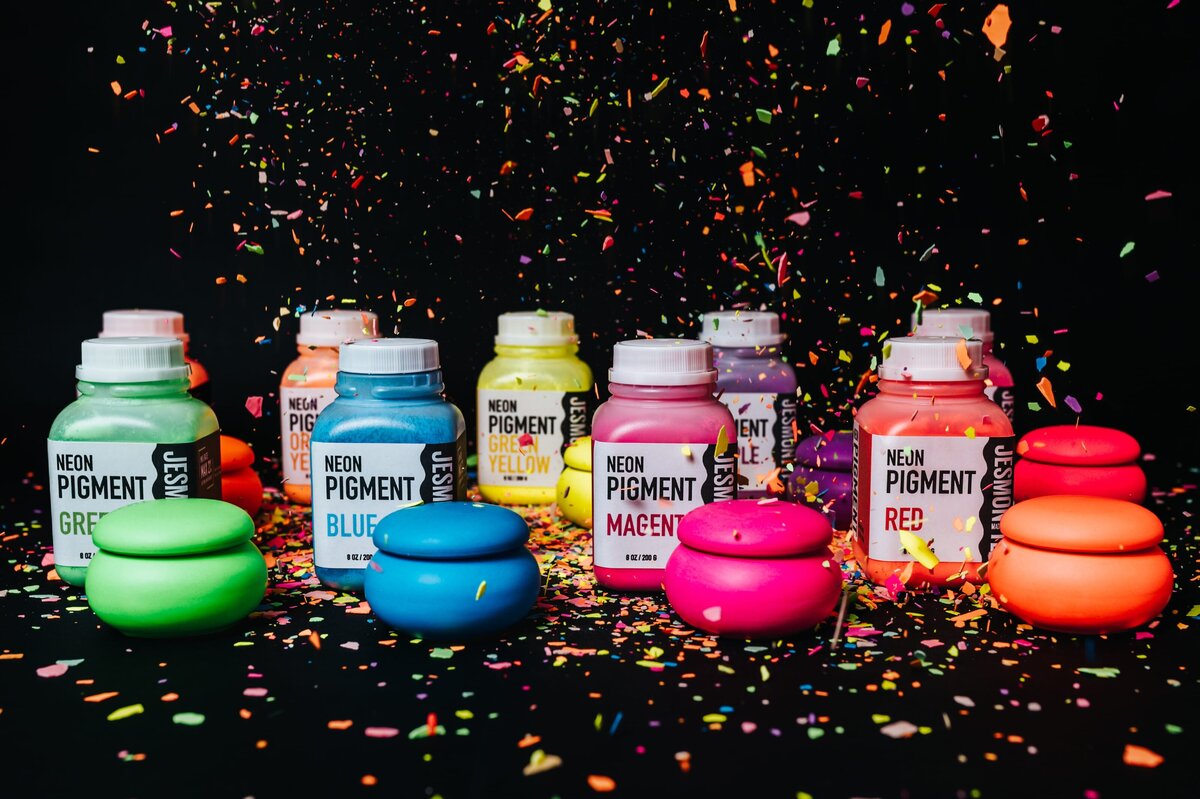 Black backdrop is splattered with vibrant paint and 9 bottles of bright paint sit on top.