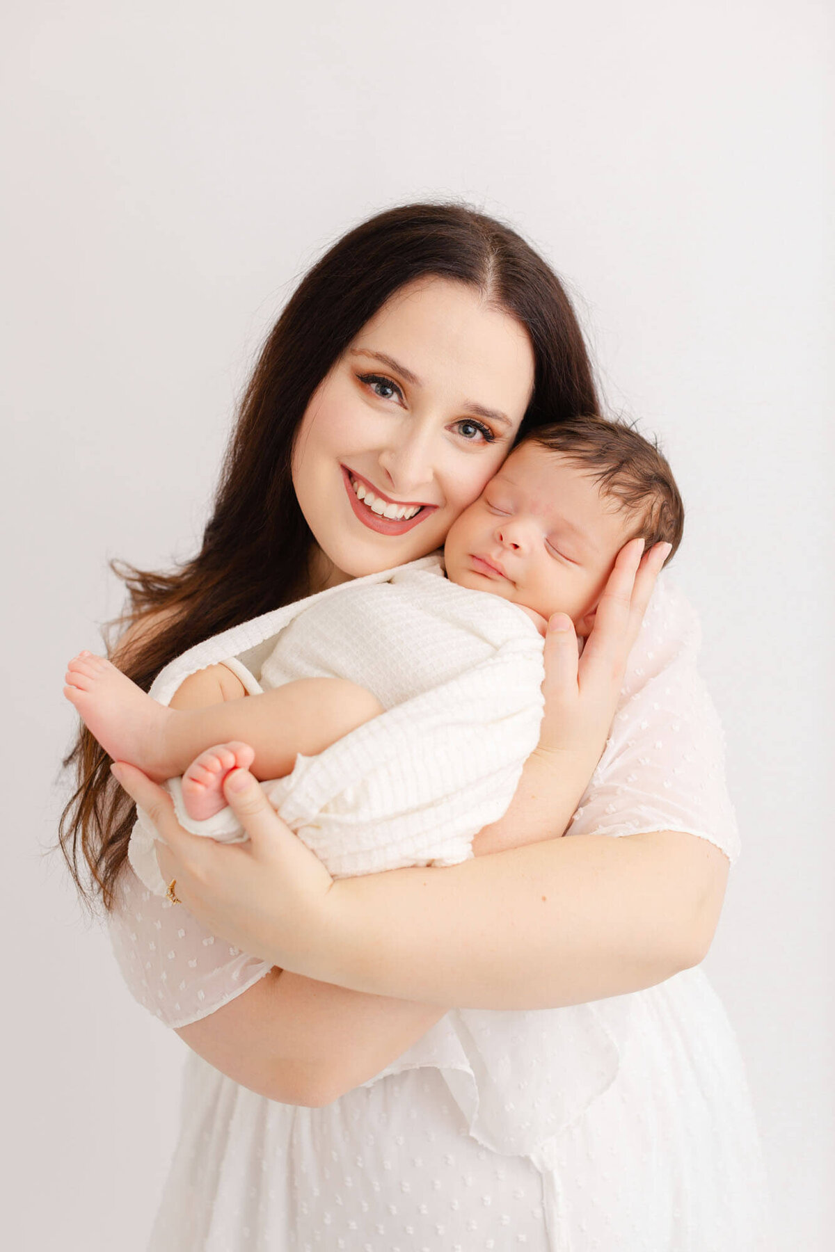 Light-skinned mom with black hair in white dress is holding newborn baby up by her cheek and smiling at the camera.
