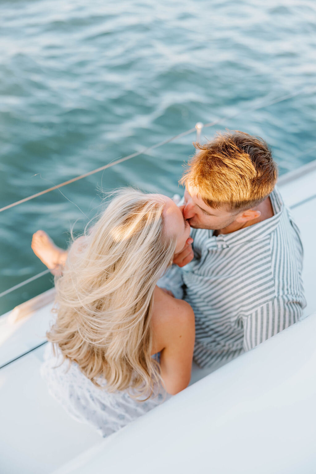 Wedding Photographer RI | A couple sits closely on a sailboat, surrounded by water. The man, in a striped shirt, kisses the smiling woman's forehead as her hair flows in the breeze.