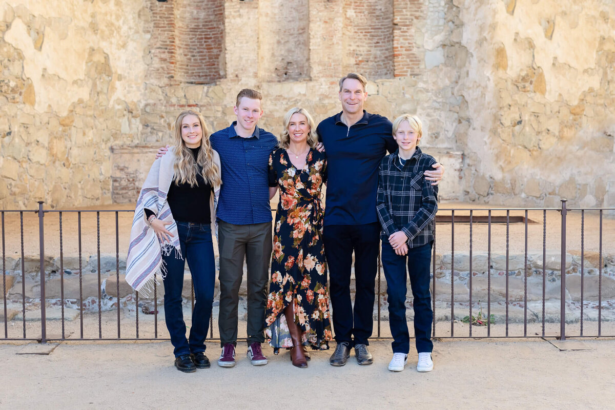A family of five wearing shades of blue, black, and orange, smiling at the camera in front of a beige brick wall.