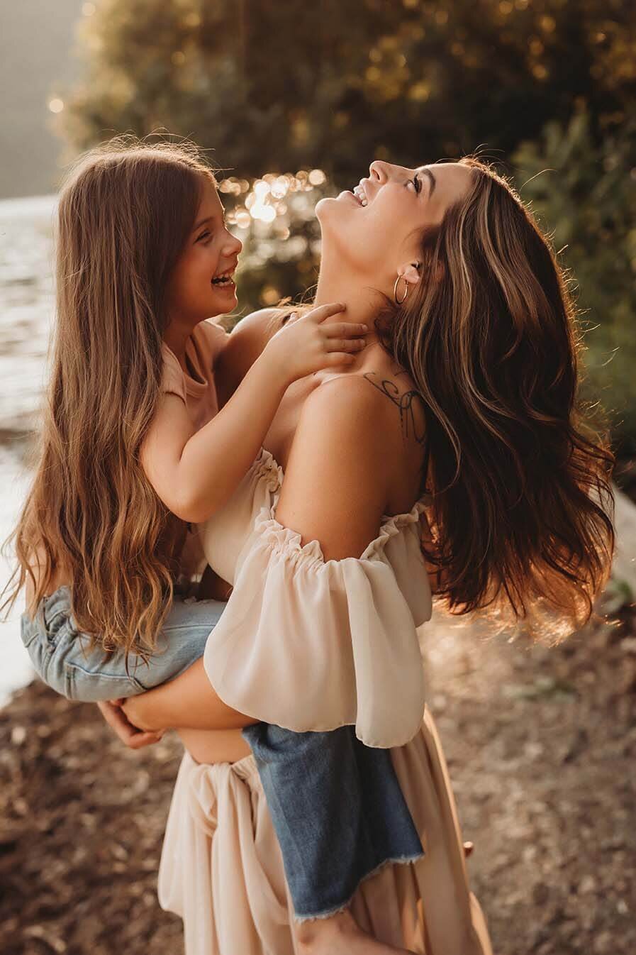 a pregnant mother holds her daughter above her belly while the daughter tickles the mothers neck. the mother throws her head back in joy and laughter