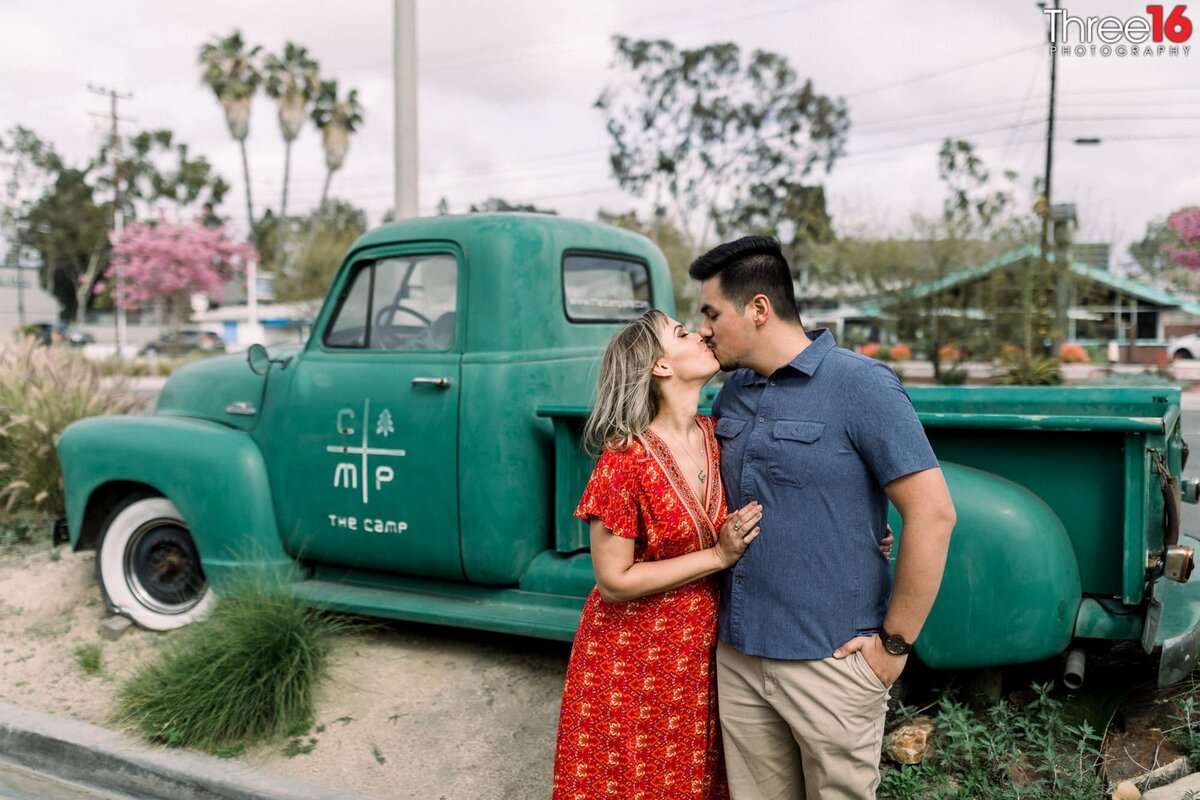 Engaged couple share a sweet kiss in front of a vintage green pickup truck