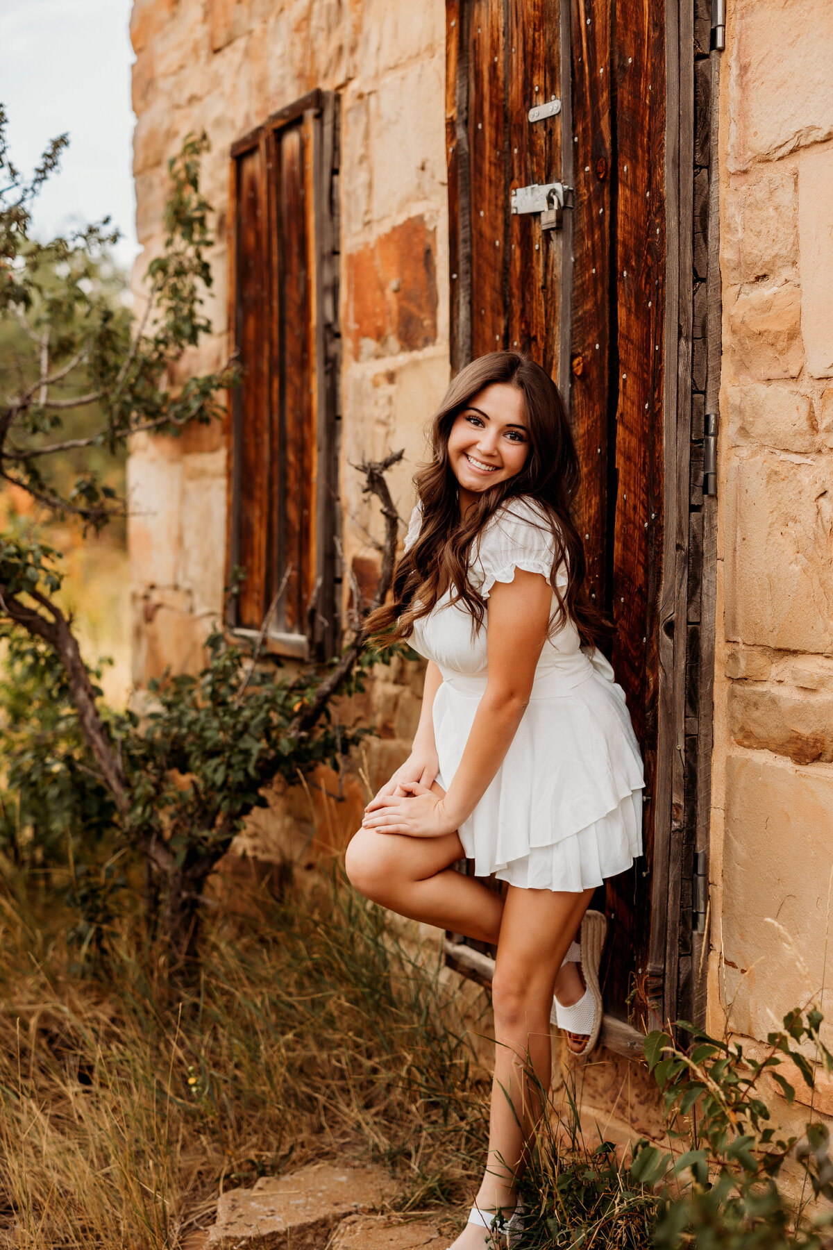 Senior girl wearing a short white romper poses against a brown barn door during her senior session in Boulder Colorado