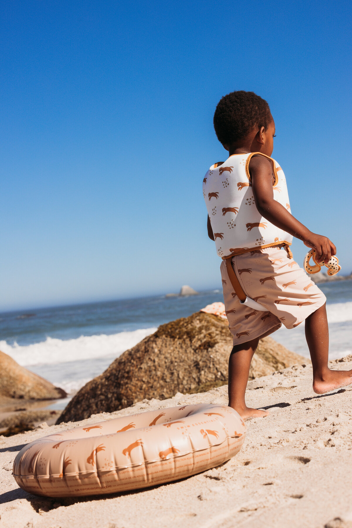 Young child wearing Swim Essentials leopard-print swim vest and sun hat, sitting on the beach while looking out at the ocean in Cape Town.