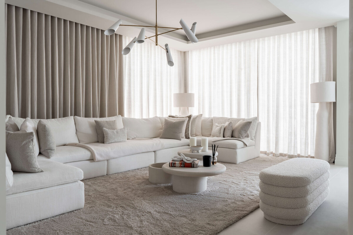 A spacious, modern living area. A large white couch with neutral throw pillows sits on a soft, textured cream rug. Behind the couch are floor-to-celiing cream drapes extending the length of the wall.