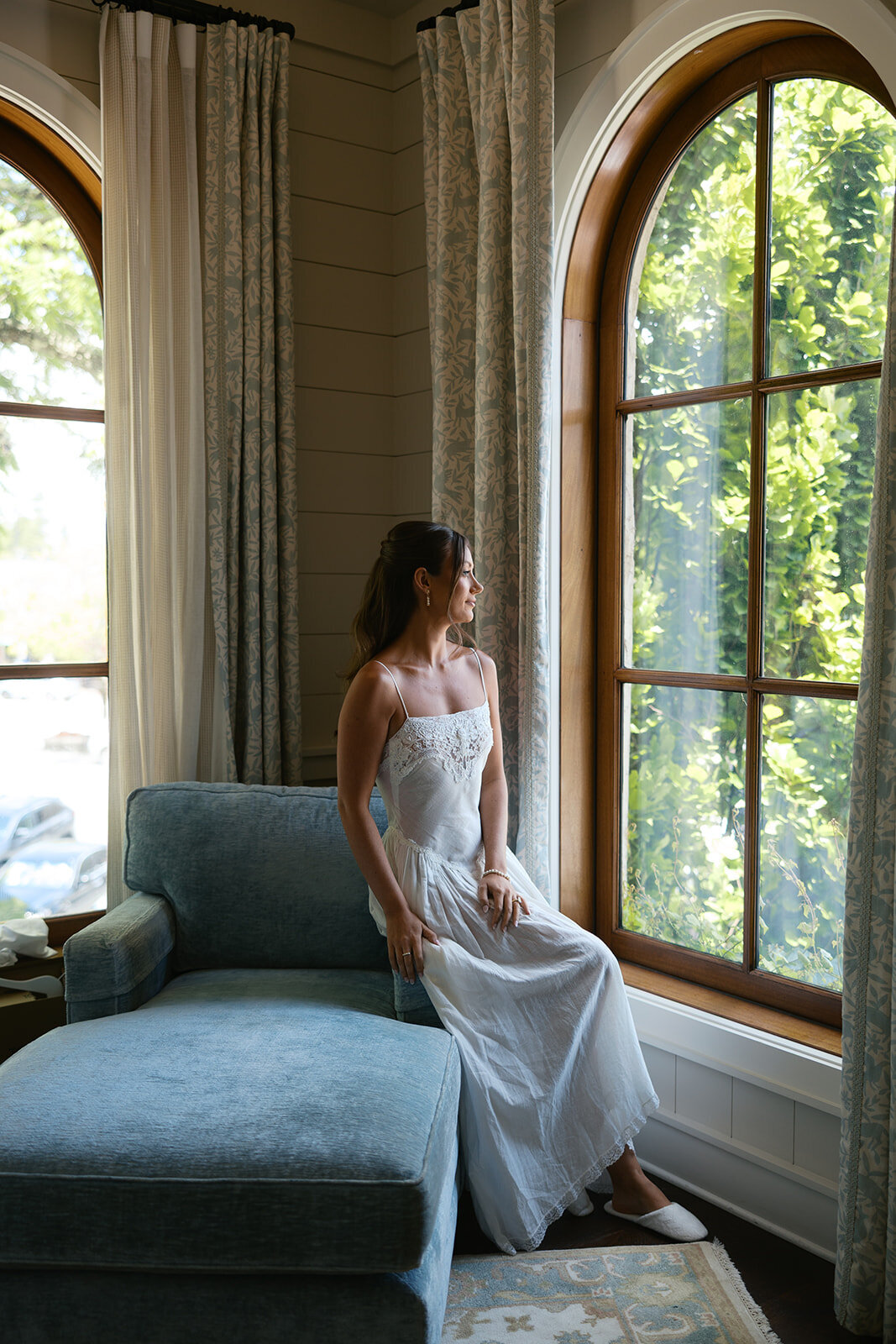 Bride in a white lace slip looks out the window in a soft blue spa suite at Old Edwards Inn.
