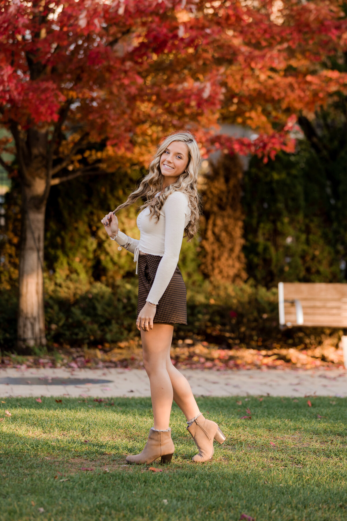 A blonde girl twirls her hair in front of red and orange colored trees for her senior photos.